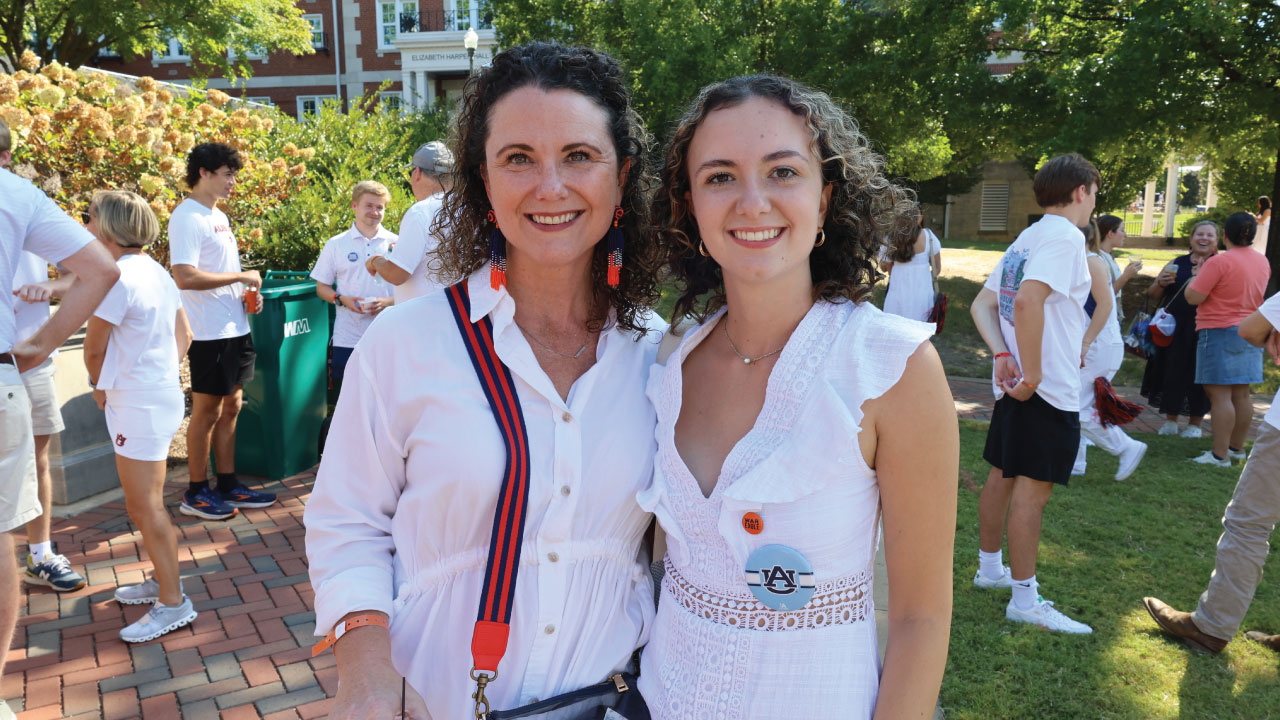 image of two women in white with auburn buttons. they are at a tailgate with activity behind them.