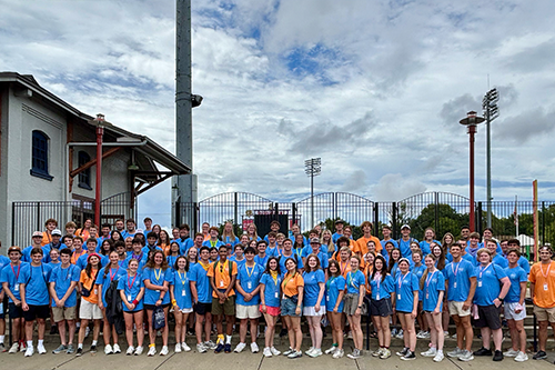 Large group of students standing together outdoors on a paved area near a gated entrance and stadium light poles, wearing mostly blue or orange shirts with lanyards under a cloudy sky.