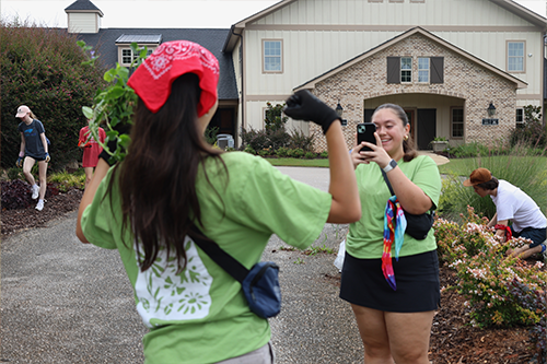One student wearing a red bandana holds a handful of plants while another student in a green shirt takes a photo with a phone in a residential neighborhood, with additional students working in a landscaped area nearby.