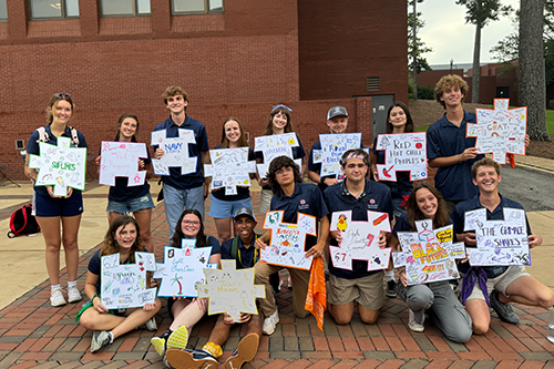 Group of students pose outdoors on a brick walkway holding handmade signs with drawings and text, standing in front of a large brick building.