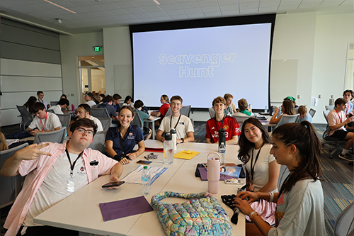 Students sit around a table in a classroom with bags, water bottles, and papers, while a large screen in the background displays the words “Scavenger Hunt.”