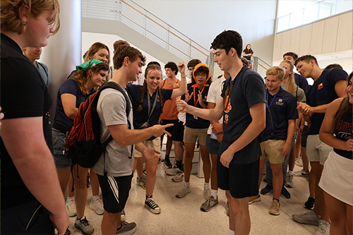 Students gather in a lobby area around two participants playing rock-paper-scissors, with others watching closely and wearing lanyards.