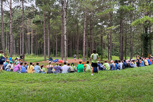 Large group of students sit in a wide circle on a grassy field surrounded by tall pine trees, with one person standing in the center leading an outdoor activity.