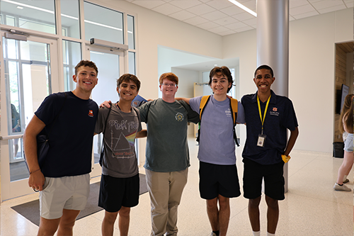 Five students stand together in a bright building lobby with glass doors and light-colored walls, posing side by side with arms around each other.