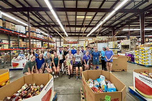 Large group of students pose inside a warehouse-style food bank or storage facility, surrounded by tall shelving, pallets of supplies, and large donation bins filled with packaged food items
