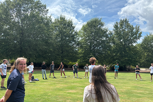Students spread out in a wide circle on a grassy field, participating in an outdoor activity surrounded by trees under a partly cloudy sky.