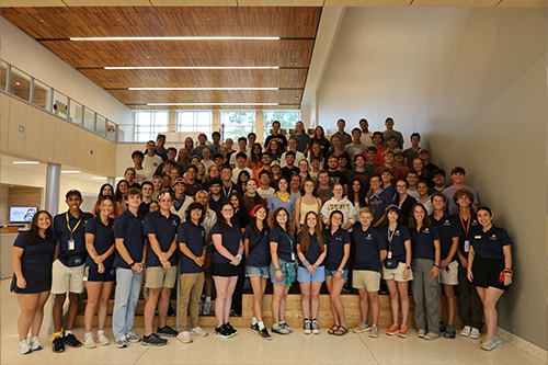 Large group of students gathered on an indoor staircase and landing in a modern building with wood-paneled ceiling and glass balcony, many wearing dark shirts and lanyards.