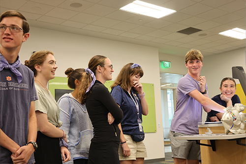 Seven students stand around a table in a classroom, some wearing lanyards, while one student works with materials on the table during a group activity.