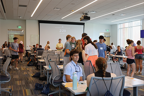 Students collaborate at tables in a large, modern classroom with rolling chairs, while others stand and talk near a projection screen displaying a timer.