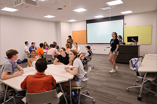 Students sit and talk in small groups around tables in a classroom, with a presenter and a projected slide visible at the front of the room.