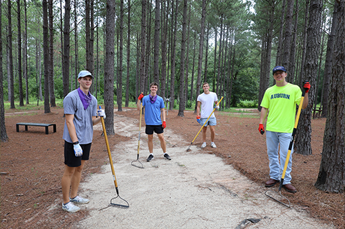 Four students stand on a dirt path in a wooded area holding landscape rakes, wearing gloves and casual clothing, with tall pine trees surrounding them.